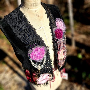 K&ouml;nnte beinhalten: Schwarze Samtweste mit schwarzer Spitzenborte, Perlen und Stoffblumen in Rosa- und Burgundert&ouml;nen. Die Weste wird auf einer Vintage-Mannequin pr&auml;sentiert, die ihre aufwendigen Details und Texturen zeigt.