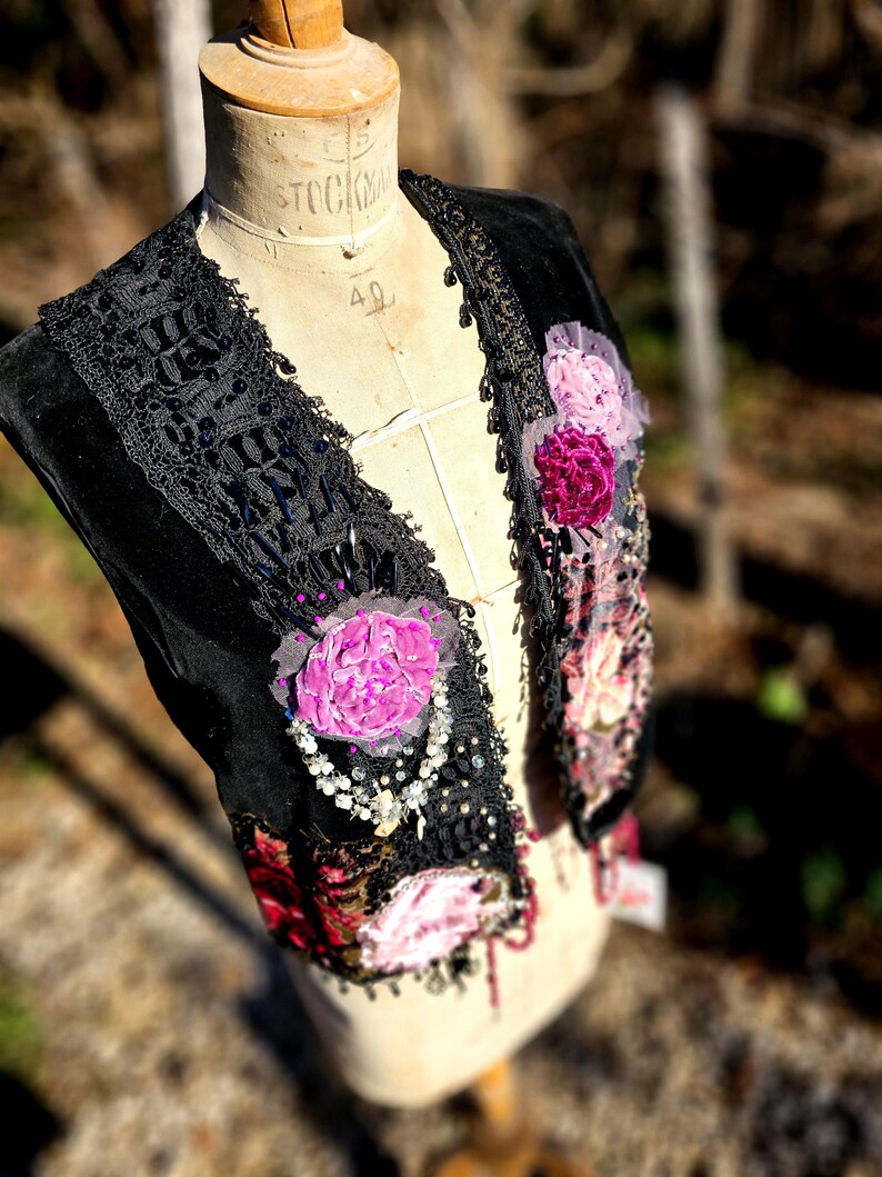 K&ouml;nnte beinhalten: Schwarze Samtweste mit schwarzer Spitzenborte, Perlen und Stoffblumen in Rosa- und Burgundert&ouml;nen. Die Weste wird auf einer Vintage-Mannequin pr&auml;sentiert, die ihre aufwendigen Details und Texturen zeigt.