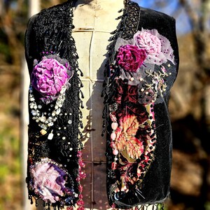 K&ouml;nnte beinhalten: Schwarze Samtweste mit aufwendigen floralen Verzierungen in Rosa- und Lilat&ouml;nen. Die Weste hat Perlenfransen, Spitzensaum und dekorative Perlenarbeiten. Die West ist auf einer Vintage-Schneiderpuppe ausgestellt.