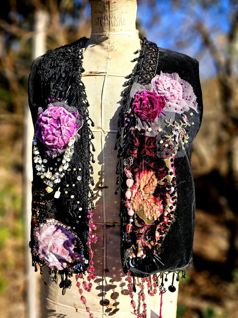 K&ouml;nnte beinhalten: Schwarze Samtweste mit aufwendigen floralen Verzierungen in Rosa- und Lilat&ouml;nen. Die Weste hat Perlenfransen, Spitzensaum und dekorative Perlenarbeiten. Die West ist auf einer Vintage-Schneiderpuppe ausgestellt.