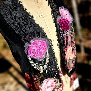 K&ouml;nnte beinhalten: Schwarze Samtweste mit aufwendiger Spitzenborte und Perlenakzenten. Die Weste hat applizierte rosa und lila Stoffblumen und eine zarte Perlenkette. Das Kleidungsst&uuml;ck wird auf einer Vintage-Schneiderpuppe pr&auml;sentiert.