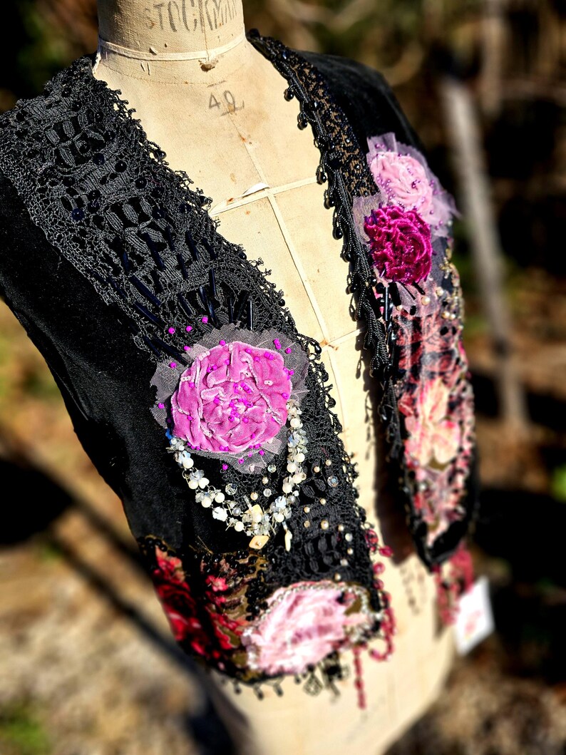 K&ouml;nnte beinhalten: Schwarze Samtweste mit aufwendiger Spitzenborte und Perlenakzenten. Die Weste hat applizierte rosa und lila Stoffblumen und eine zarte Perlenkette. Das Kleidungsst&uuml;ck wird auf einer Vintage-Schneiderpuppe pr&auml;sentiert.