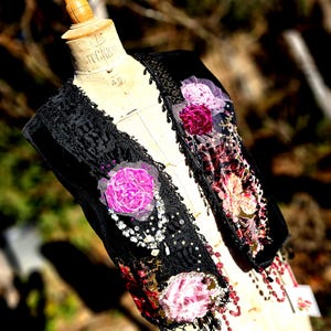 K&ouml;nnte beinhalten: Eine schwarze Samtweste mit aufwendiger Spitzenborte und floralen Verzierungen. Die Weste hat rosa und bordeauxfarbene Stoffblumen, Perlenakzente und eine dekorative Bord&uuml;re. Das Kleidungsst&uuml;ck wird auf einer Vintage-Schneiderpuppe pr&auml;sentiert.