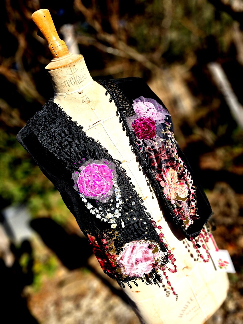 K&ouml;nnte beinhalten: Eine schwarze Samtweste mit aufwendiger Spitzenborte und floralen Verzierungen. Die Weste hat rosa und bordeauxfarbene Stoffblumen, Perlenakzente und eine dekorative Bord&uuml;re. Das Kleidungsst&uuml;ck wird auf einer Vintage-Schneiderpuppe pr&auml;sentiert.