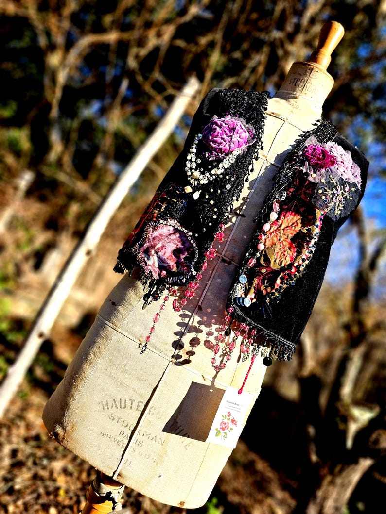 K&ouml;nnte beinhalten: Eine schwarze Weste mit floralen Verzierungen, Perlen und Spitze, pr&auml;sentiert auf einer Vintage-Schneiderpuppe. Die Weste hat lila und rosa Blumen mit herabh&auml;ngenden Perlenakzenten. Die Schneiderpuppe tr&auml;gt den Text "HAUTE COUTURE STOLKMAN PARIS BREVETE".