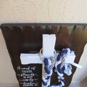 May include: A white wooden cross mounted on a dark brown wooden plaque with rough edges. A silver metal ring holds three braided ropes in navy blue, white, and silver. The text A cord of THREE strands is not easily broken ECCLESIASTES 4:12 is printed.
