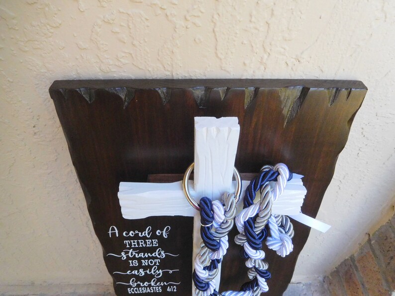 May include: A white wooden cross mounted on a dark brown wooden plaque with rough edges. A silver metal ring holds three braided ropes in navy blue, white, and silver. The text A cord of THREE strands is not easily broken ECCLESIASTES 4:12 is printed.