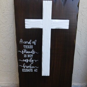 May include: A dark brown wooden wall plaque with a textured live edge, featuring a white carved wooden cross mounted in the center. The text "A cord of THREE strands is not easily broken ECCLESIASTES 4:12" is printed in white on the left side.