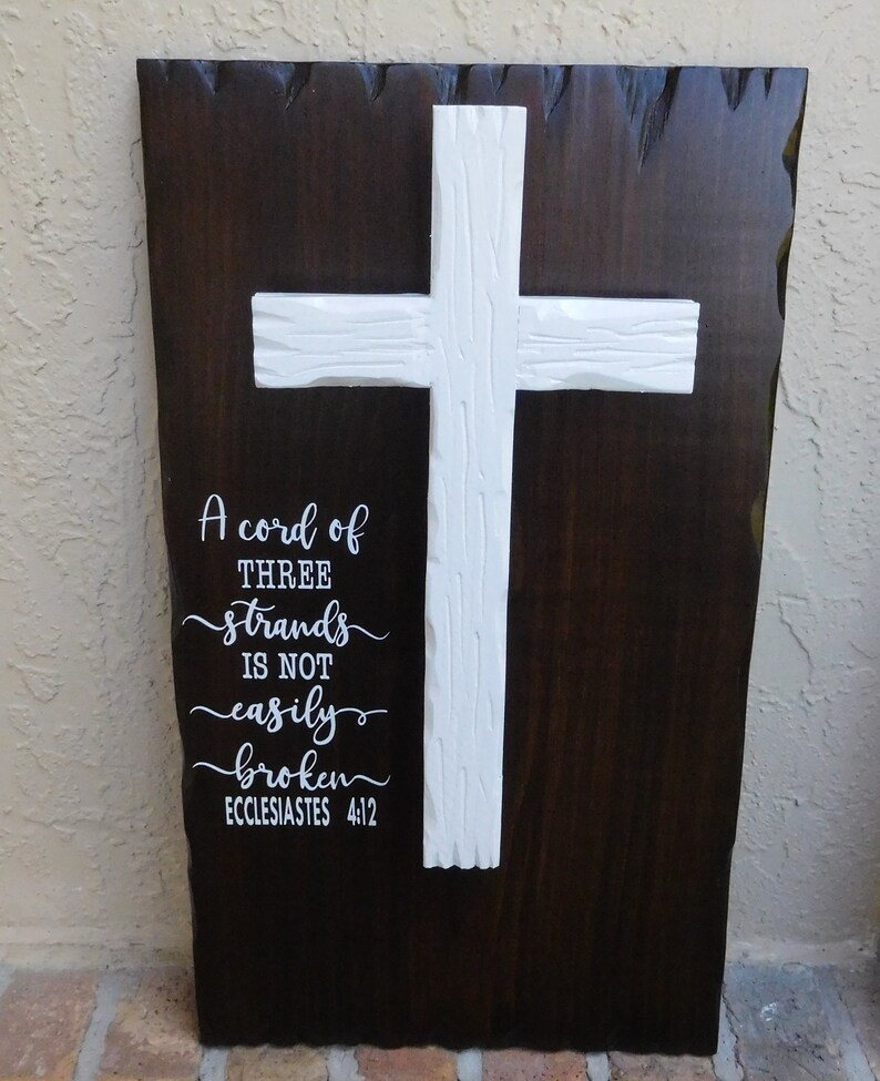 May include: A dark brown wooden wall plaque with a textured live edge, featuring a white carved wooden cross mounted in the center. The text "A cord of THREE strands is not easily broken ECCLESIASTES 4:12" is printed in white on the left side.