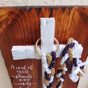 Peut inclure: Une plaque murale en bois avec une planche brun fonc&eacute; &agrave; bord brut et une croix blanche. Un anneau dor&eacute; retient trois cordes tress&eacute;es violettes, dor&eacute;es et blanches. Le texte indique : A cord of three strands is not easily broken.
