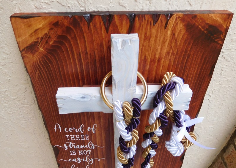 Peut inclure: Une plaque murale en bois avec une planche brun fonc&eacute; &agrave; bord brut et une croix blanche. Un anneau dor&eacute; retient trois cordes tress&eacute;es violettes, dor&eacute;es et blanches. Le texte indique : A cord of three strands is not easily broken.