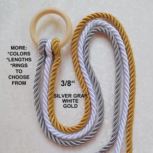 Op de afbeelding: Drie strengen touw, in zilvergrijs, wit en goud, zijn door een houten ring gelust. Het touw is 3/8 inch dik. De tekst "MORE: *COLORS *LENGTHS *RINGS TO CHOOSE FROM" is zichtbaar in de afbeelding.