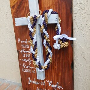 Peut inclure: Une plaque en bois rectangulaire avec une finition teint&eacute;e fonc&eacute;e pr&eacute;sente une croix peinte en blanc. Des cordes tress&eacute;es violettes, blanches et dor&eacute;es pendent d'un anneau dor&eacute; sur la croix. Le texte blanc indique : A cord of three strands is not easily broken, Jordan and Kayla, July 10, 2021.