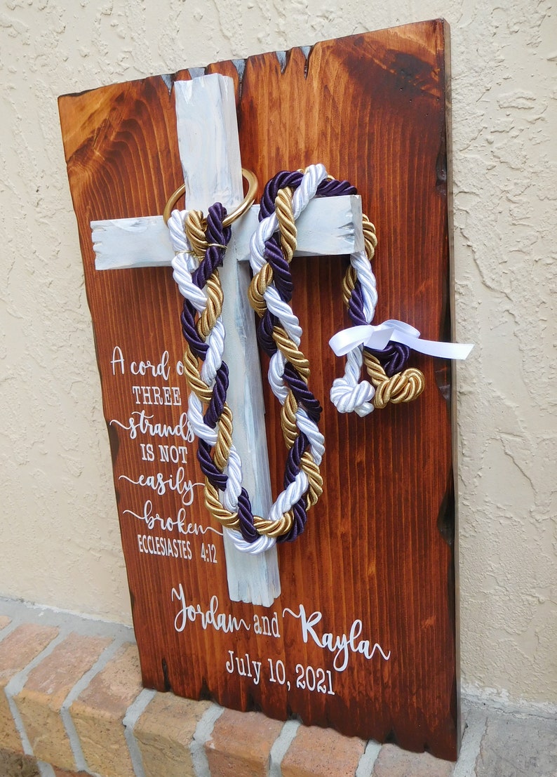 Peut inclure: Une plaque en bois rectangulaire avec une finition teint&eacute;e fonc&eacute;e pr&eacute;sente une croix peinte en blanc. Des cordes tress&eacute;es violettes, blanches et dor&eacute;es pendent d'un anneau dor&eacute; sur la croix. Le texte blanc indique : A cord of three strands is not easily broken, Jordan and Kayla, July 10, 2021.