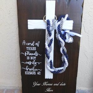 May include: A dark brown wooden plaque with a white cross mounted in the center. Three braided cords in navy blue, silver, and white hang from a metal ring on the cross. White text reads: A cord of THREE strands IS NOT easily broken ECCLESIASTES 4:12.
