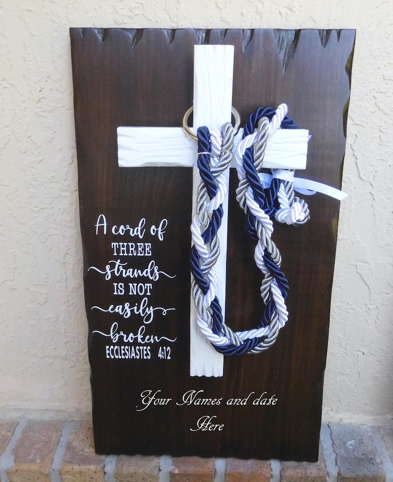 May include: A dark brown wooden plaque with a white cross mounted in the center. Three braided cords in navy blue, silver, and white hang from a metal ring on the cross. White text reads: A cord of THREE strands IS NOT easily broken ECCLESIASTES 4:12.
