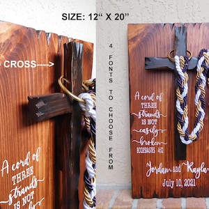 May include: A rectangular wooden wall plaque measuring 30.48 cm by 50.8 cm, featuring a dark brown 3D cross with a gold ring and braided purple, gold, and white cords. White text reads: A cord of three strands is not easily broken, Ecclesiastes 4:12, Jordam and Kaylar, July 10, 2021.