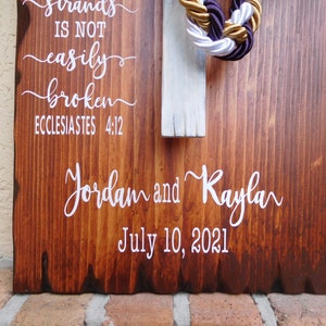 Peut inclure: Une plaque en bois rectangulaire avec une finition teint&eacute;e brun fonc&eacute; et des bords naturels. Elle pr&eacute;sente un texte blanc indiquant "strands IS NOT easily broken ECCLESIASTES 4:12", "Jordam and Kayla" et "July 10, 2021". Une croix blanche avec des cordes tress&eacute;es dor&eacute;es, blanches et violettes.