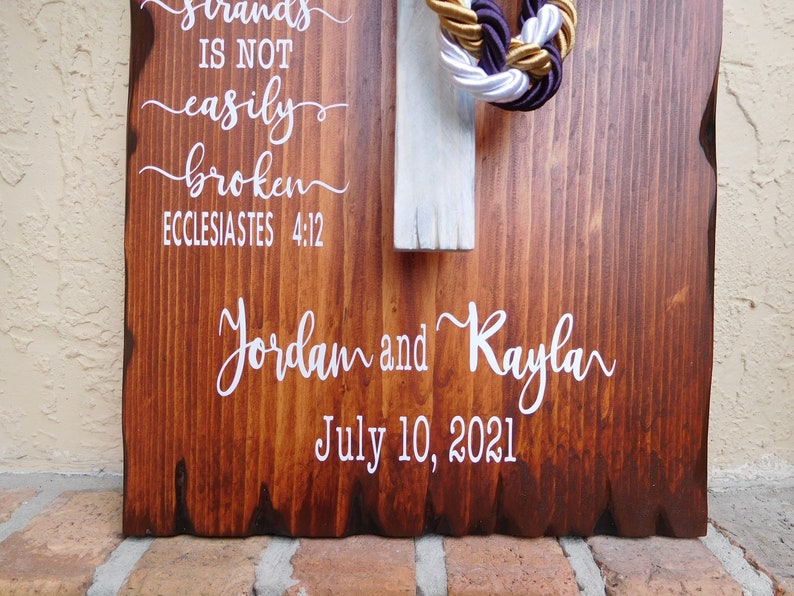 Peut inclure: Une plaque en bois rectangulaire avec une finition teint&eacute;e brun fonc&eacute; et des bords naturels. Elle pr&eacute;sente un texte blanc indiquant "strands IS NOT easily broken ECCLESIASTES 4:12", "Jordam and Kayla" et "July 10, 2021". Une croix blanche avec des cordes tress&eacute;es dor&eacute;es, blanches et violettes.