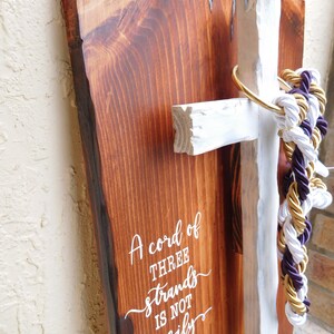 Peut inclure: Une plaque en bois rectangulaire &agrave; bord brut avec une finition teint&eacute;e brun fonc&eacute; portant le texte blanc A cord of THREE strands IS NOT easily broken ECCLESIASTES 4:12. Une croix en bois peinte en blanc est fix&eacute;e avec un anneau dor&eacute; et des cordes.