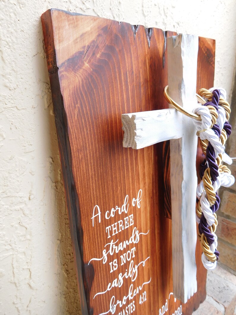 Peut inclure: Une plaque en bois rectangulaire &agrave; bord brut avec une finition teint&eacute;e brun fonc&eacute; portant le texte blanc A cord of THREE strands IS NOT easily broken ECCLESIASTES 4:12. Une croix en bois peinte en blanc est fix&eacute;e avec un anneau dor&eacute; et des cordes.