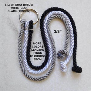 May include: A decorative wedding unity cord set with three twisted ropes in silver gray, white, and black, attached to a gold ring. The text on the image says "SILVER GRAY (BRIDE), WHITE (GOD), BLACK (GROOM)" and "3/8". Additional text reads "MORE: COLORS, LENGTHS, RINGS TO CHOOSE FROM".