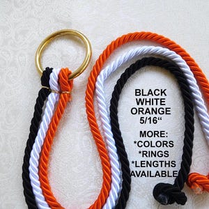 May include: A gold-toned metal ring attached to three twisted ropes in black, white, and orange. The text on the image reads: BLACK WHITE ORANGE 5/16" MORE: *COLORS *RINGS *LENGTHS AVAILABLE. The ropes are knotted at the ends and hang downwards.