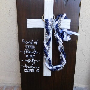 May include: A dark brown wooden plaque with a white cross mounted on it. A braided cord in navy blue, silver, and white hangs from a gold ring on the cross. The text reads: A cord of THREE strands IS NOT easily broken ECCLESIASTES 4:12.