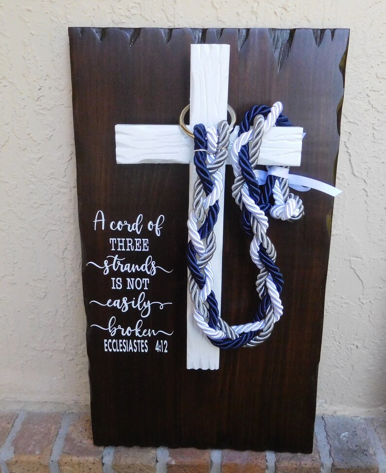 May include: A dark brown wooden plaque with a white cross mounted on it. A braided cord in navy blue, silver, and white hangs from a gold ring on the cross. The text reads: A cord of THREE strands IS NOT easily broken ECCLESIASTES 4:12.