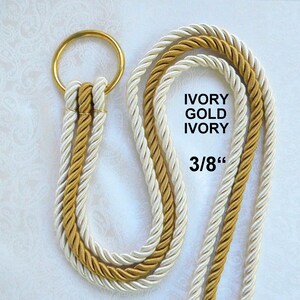 Peut inclure: Un ensemble de cordons d&eacute;coratifs compos&eacute; de deux brins torsad&eacute;s ivoire et d'un brin dor&eacute;, fix&eacute;s &agrave; un anneau m&eacute;tallique dor&eacute;. Le texte IVORY GOLD IVORY et 3/8" est imprim&eacute; sur l'image, indiquant le motif de couleur et le diam&egrave;tre de 3/8 pouce.