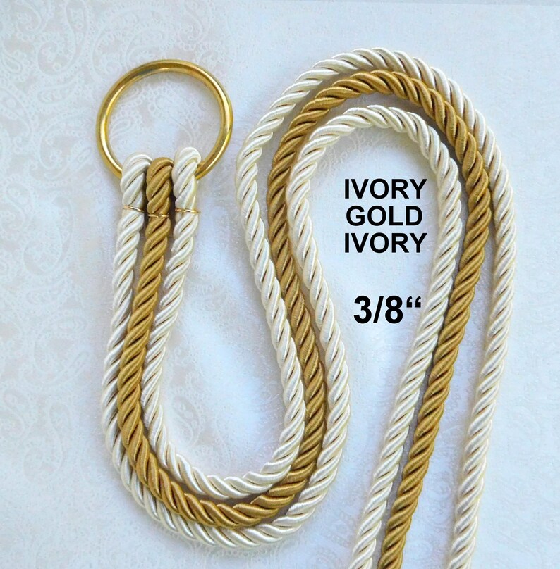 Peut inclure: Un ensemble de cordons d&eacute;coratifs compos&eacute; de deux brins torsad&eacute;s ivoire et d'un brin dor&eacute;, fix&eacute;s &agrave; un anneau m&eacute;tallique dor&eacute;. Le texte IVORY GOLD IVORY et 3/8" est imprim&eacute; sur l'image, indiquant le motif de couleur et le diam&egrave;tre de 3/8 pouce.