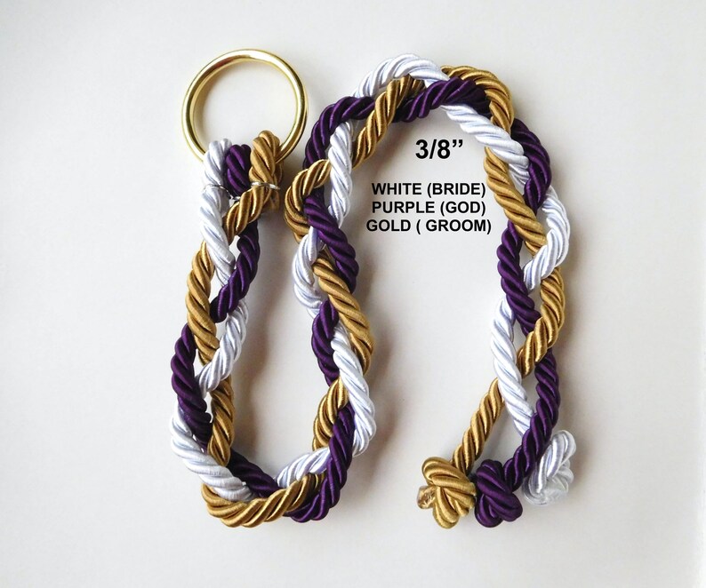 May include: A braided rope cord featuring three strands in white, purple, and gold, attached to a gold-colored metal ring. The text 3/8&rdquo;, WHITE (BRIDE), PURPLE (GOD), GOLD (GROOM) is printed on the image, indicating a 3/8 inch diameter.