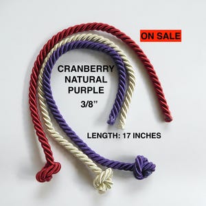 Op de afbeelding: Drie spoelen koord, een rode, een crème en een paarse, elk met een knoop vastgemaakt. Het koord is 3/8 inch dik en 17 inch lang. De tekst "Cranberry Natural Purple 3/8" en "LENGTH: 17 INCHES" is gedrukt op een witte achtergrond. De tekst "ON SALE" is gedrukt in rood op een witte achtergrond.