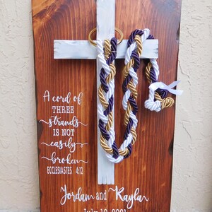 Peut inclure: Une plaque rectangulaire en bois avec une finition teint&eacute;e fonc&eacute;e et une croix peinte en blanc. Trois cordons tress&eacute;s violet, blanc et dor&eacute; pendent d'un anneau dor&eacute; sur la croix. Le texte blanc indique : A cord of THREE strands IS NOT easily broken ECCLESIASTES 4:12, Jordan and Kayla, July 10, 2021.