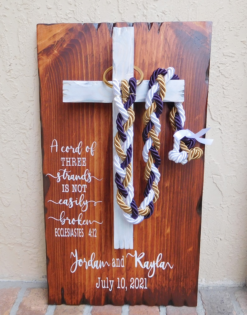 Peut inclure: Une plaque rectangulaire en bois avec une finition teint&eacute;e fonc&eacute;e et une croix peinte en blanc. Trois cordons tress&eacute;s violet, blanc et dor&eacute; pendent d'un anneau dor&eacute; sur la croix. Le texte blanc indique : A cord of THREE strands IS NOT easily broken ECCLESIASTES 4:12, Jordan and Kayla, July 10, 2021.