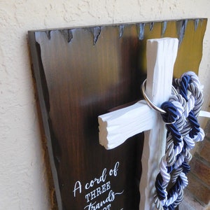 May include: A dark brown wooden plaque with a white cross attached to the front. A silver metal ring holds three braided ropes in navy blue, silver, and white. The plaque features white text that reads A cord of THREE strands is not easily broken ECCLESIASTES 4:12.