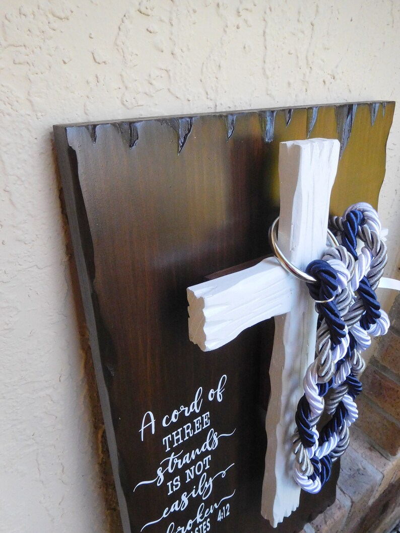 May include: A dark brown wooden plaque with a white cross attached to the front. A silver metal ring holds three braided ropes in navy blue, silver, and white. The plaque features white text that reads A cord of THREE strands is not easily broken ECCLESIASTES 4:12.