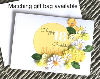 May include: A white birthday card with a yellow circle featuring the text Happy 18th Birthday Maisie. The card is decorated with layered paper flowers in shades of yellow and white, green paper leaves, and small clear rhinestones.