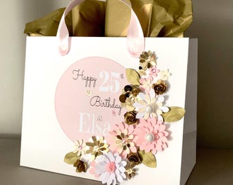 May include: A white gift bag with pink ribbon handles and gold tissue paper. A pink circle on the front features the text Happy 25th Birthday Elsa. The bag is decorated with layered paper flowers in shades of pink, white, and metallic gold.