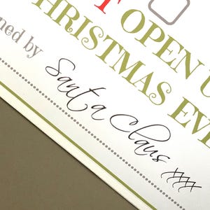 May include: A white card with text in various fonts and colors. The text includes "OPEN UP CHRISTMAS EVE" in green and red, and "Signed by Santa Claus" in black script. A dotted line and green border frame the text.