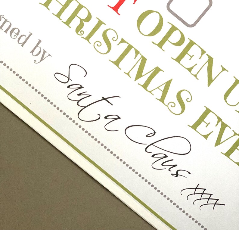 May include: A white card with text in various fonts and colors. The text includes "OPEN UP CHRISTMAS EVE" in green and red, and "Signed by Santa Claus" in black script. A dotted line and green border frame the text.