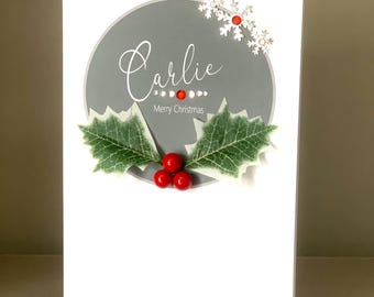 May include: A white greeting card featuring a gray circle with the name Carlie and the text Merry Christmas. The design includes three red berries, two green holly leaves, silver snowflake accents, and small red and clear crystal embellishments.