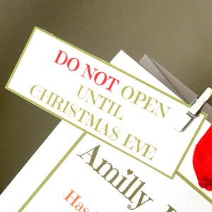 May include: A white rectangular gift tag with the text "DO NOT OPEN UNTIL CHRISTMAS EVE" in red and green lettering. The tag is clipped to a gift with a silver clip. The word "Amilly" is also visible on a lower tag.