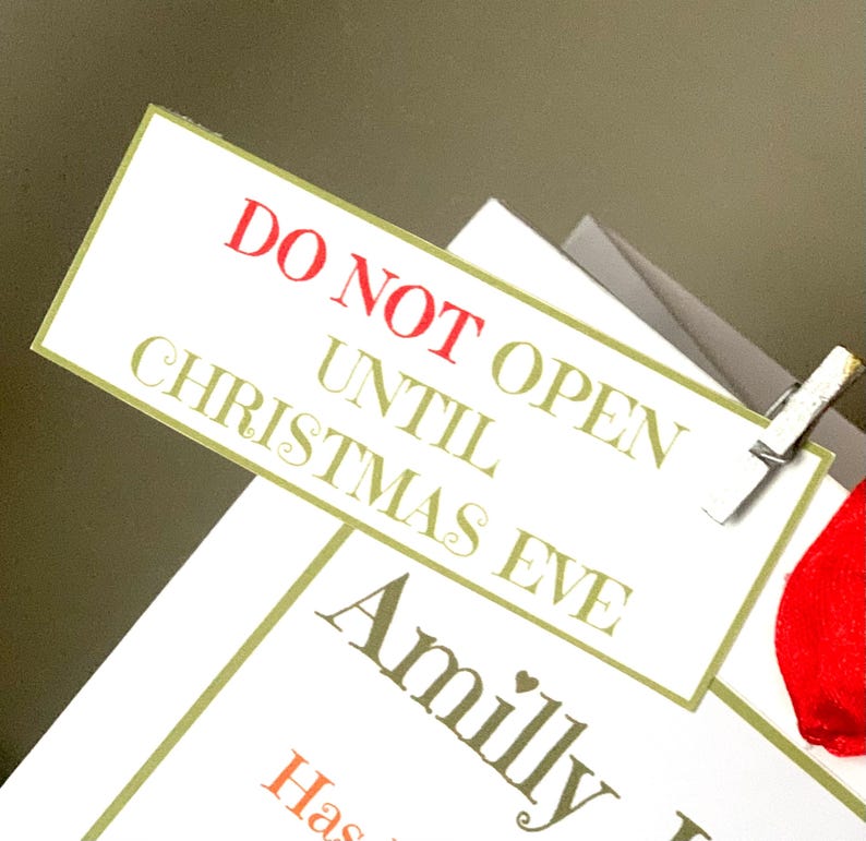 May include: A white rectangular gift tag with the text "DO NOT OPEN UNTIL CHRISTMAS EVE" in red and green lettering. The tag is clipped to a gift with a silver clip. The word "Amilly" is also visible on a lower tag.