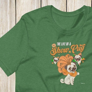 May include: Green t-shirt featuring a cartoon pug wearing a lion's mane and the text "The Life of a Show Pug." The design includes additional pug illustrations. The shirt is made of a soft, comfortable material.