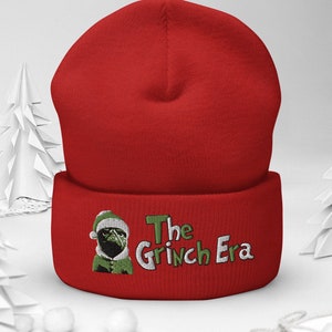 Może przedstawiać: Czerwona czapka z haftowanym napisem "The Grinch Era". Czapka posiada haftowany wizerunek mopsa w czapce Świętego Mikołaja i zielonym stroju. Czapka jest prezentowana na białej powierzchni z papierowymi choinkami.