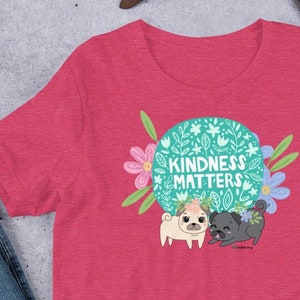 May include: A pink t-shirt with a green circle design featuring two pugs and the text "Kindness Matters". The design includes flowers and leaves.
