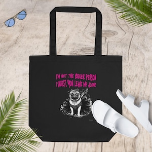 May include: A black tote bag featuring a graphic of a pug wearing a black tutu and a spiked collar. Above the dog, the text reads in bright pink letters: I'M NOT THE BIGGER PERSON I SUGGEST YOU LEAVE ME ALONE. The bag has two black shoulder straps.
