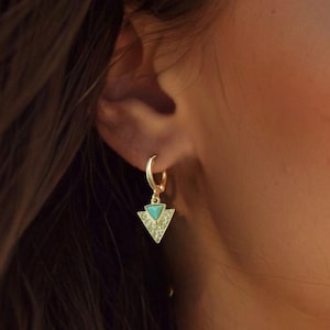 May include: Gold hoop earrings with a triangular pendant. The pendant features a turquoise-colored stone at the top and is surrounded by small, clear crystals. The earrings are a delicate piece of jewelry.