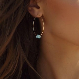 May include: A close-up shot of a person wearing a gold hoop earring with a small, light blue bead. The person is wearing a white halter top, and the background is a blurred view of the ocean and sky.